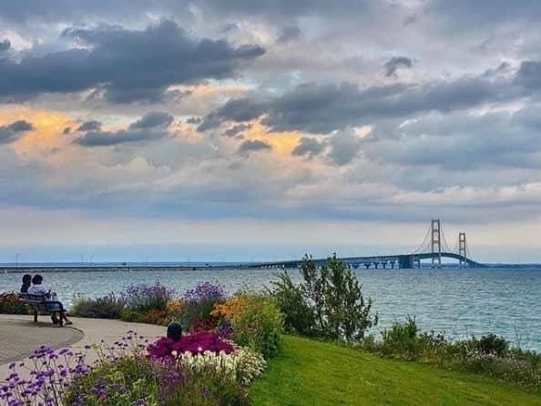 The Mackinac Bridge at dusk seen across the water from Bridge View Park, with a flower garden and a couple seated on a bench in the foreground