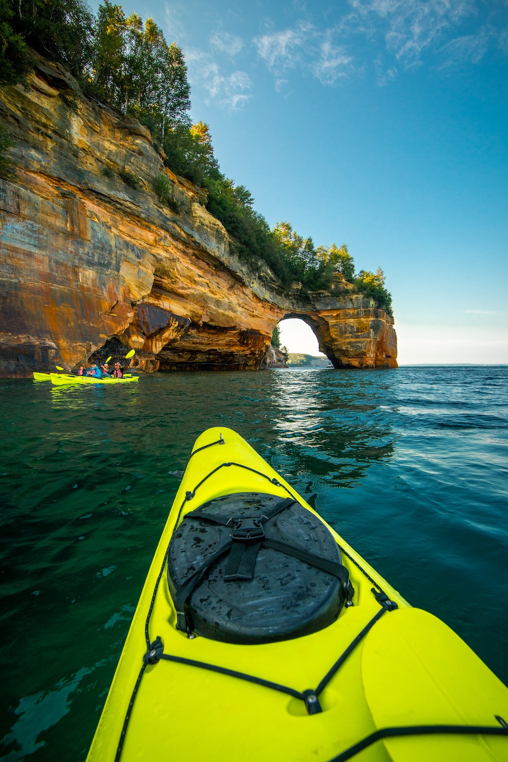 What Do I Do When I Get to Munising? View over the bow of a yellow sea kayak toward a sandstone arch at Pictured Rocks, with a tandem kayak of paddlers alongside the cliff