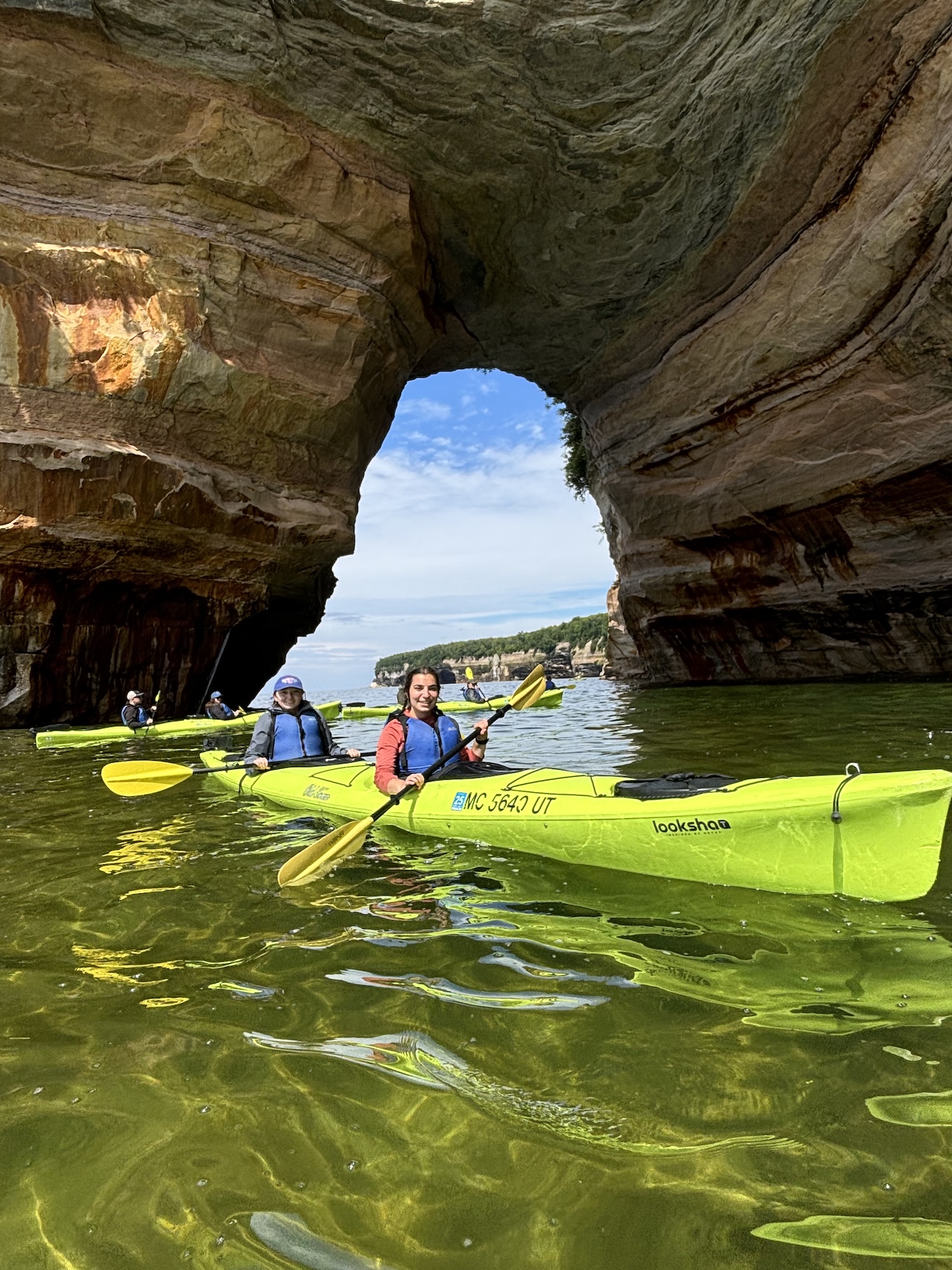 Pictured Rocks National Lakeshore Kayakers in a green tandem kayak paddling inside a large sandstone sea arch at Pictured Rocks National Lakeshore, with blue sky visible through the opening